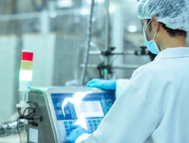 AI training food manufacturing: Masked operator in a hairnet and blue gloves using a touchscreen control panel on a food production machine.