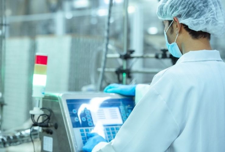 AI training food manufacturing: Masked operator in a hairnet and blue gloves using a touchscreen control panel on a food production machine.