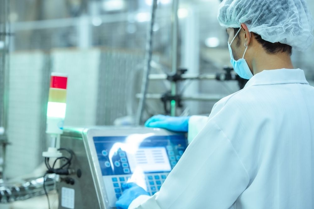 AI training food manufacturing: Masked operator in a hairnet and blue gloves using a touchscreen control panel on a food production machine.