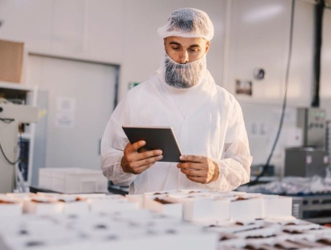 Continuous Learning in Food Manufacturing: A bearded man in a white hairnet and food production overalls holding a tablet while checking rows of packaged chocolate products on a factory production line.