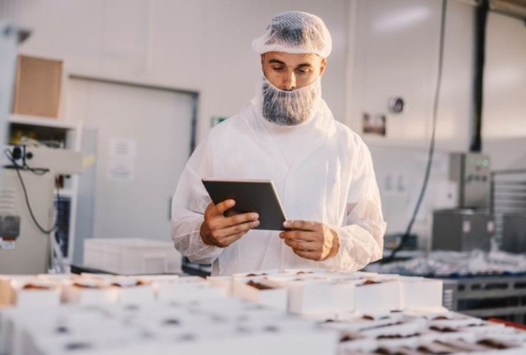 Continuous Learning in Food Manufacturing: A bearded man in a white hairnet and food production overalls holding a tablet while checking rows of packaged chocolate products on a factory production line.