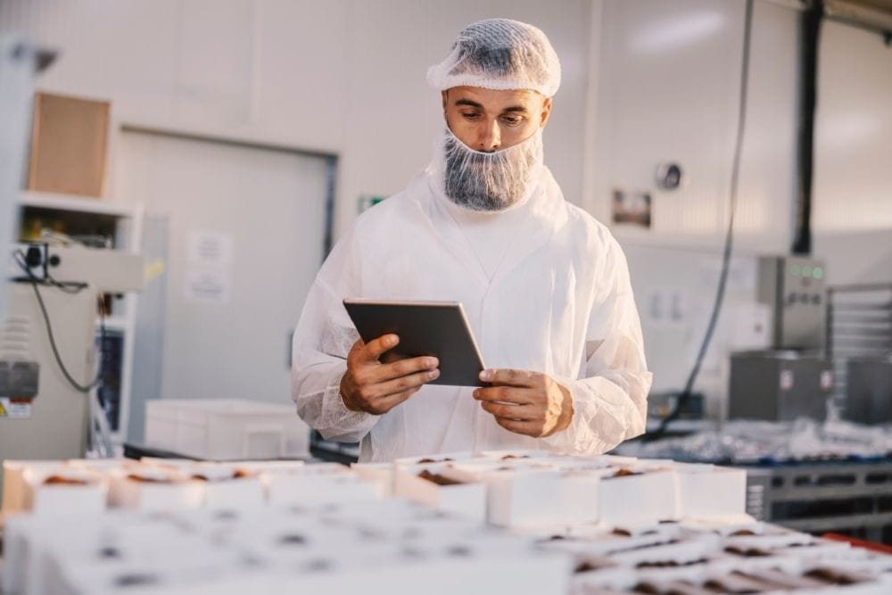 Continuous Learning in Food Manufacturing: A bearded man in a white hairnet and food production overalls holding a tablet while checking rows of packaged chocolate products on a factory production line.