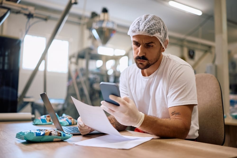 Food factory online training: Food factory worker wearing protective clothing, reviewing paperwork and a mobile phone at a desk inside a production facility.