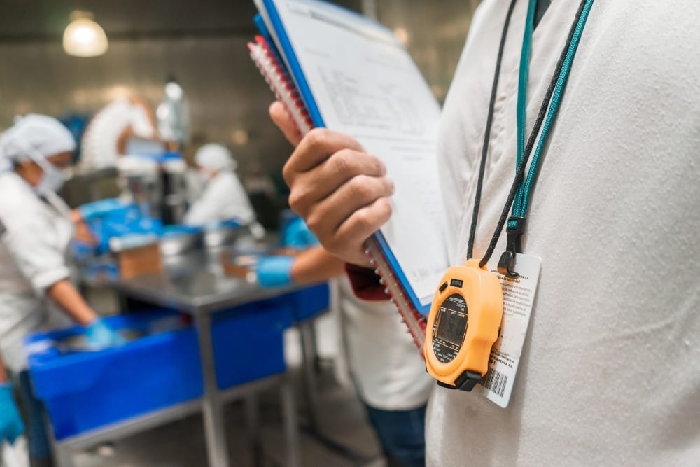 Food factory quality inspection: Supervisor holding a clipboard and stopwatch while observing food production staff working on a factory line.