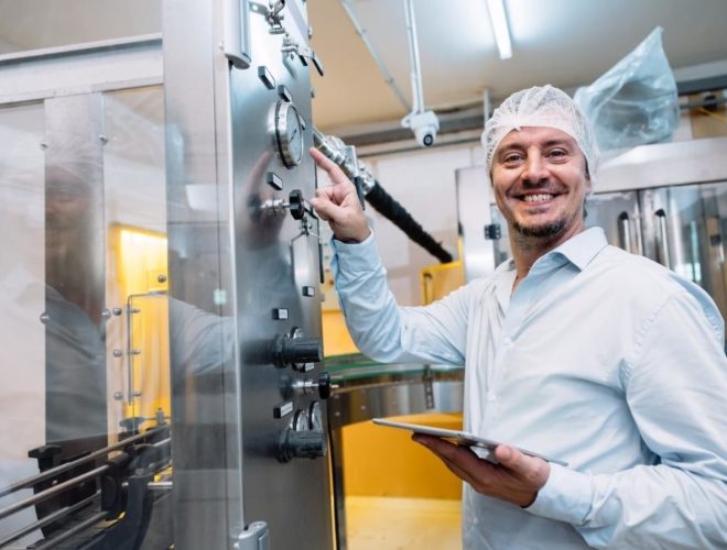 How to reduce turnover on food production lines: A smiling male food factory worker in a hairnet and white shirt holding a tablet whilst adjusting controls on a stainless steel production machine.
