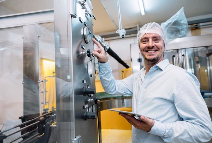 How to reduce turnover on food production lines: A smiling male food factory worker in a hairnet and white shirt holding a tablet whilst adjusting controls on a stainless steel production machine.