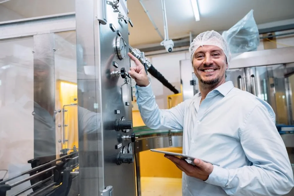 How to reduce turnover on food production lines: A smiling male food factory worker in a hairnet and white shirt holding a tablet whilst adjusting controls on a stainless steel production machine.