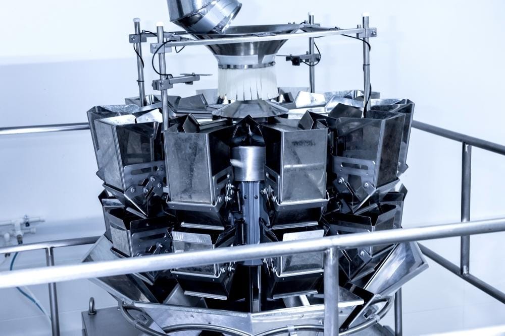 Bitesize learning in food production Close-up of a stainless steel multihead weigher machine used in food production, with multiple weighing buckets arranged in a circular formation.
