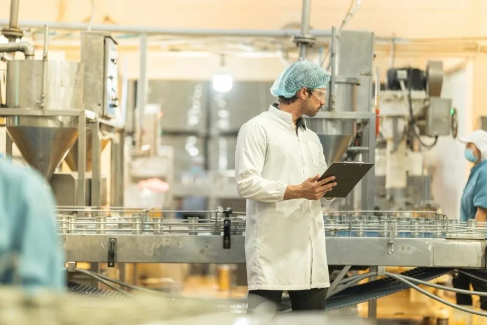 food processing efficiency: A food factory worker in a white coat, hair net and safety glasses reviewing a clipboard beside a bottling conveyor line, with other workers visible in the background.