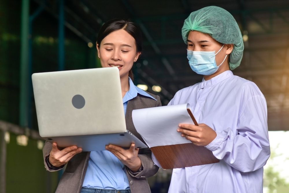 future of food manufacturing training: Office worker with a laptop and a masked food factory worker with a clipboard reviewing information together.