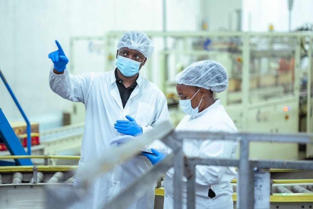 how do food manufacturers retain operator knowledge on the line: A male supervisor in a hairnet, face mask and gloves pointing and speaking to a female colleague holding a clipboard on a food production line, with packaging machinery in the background.