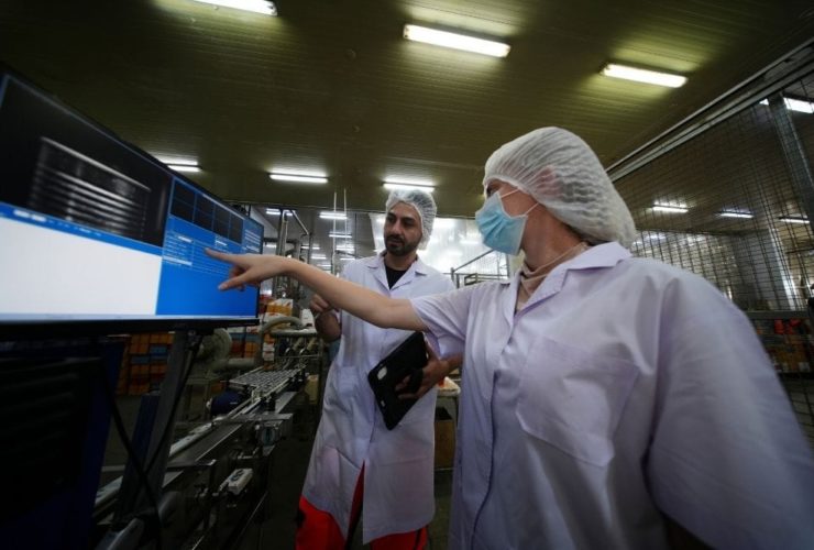 How does faster knowledge access reduce food production downtime: Two food production workers in white coats and hairnets reviewing data on a wall-mounted monitor on the factory floor.