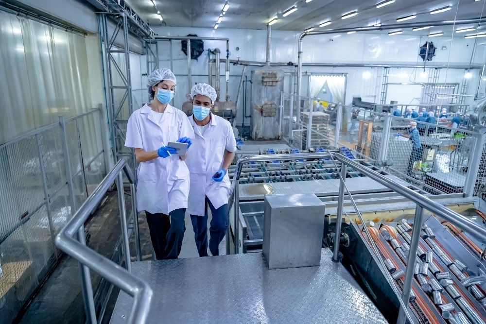 how is AI changing frontline training in food production: Two workers in hairnets, masks and white coats reviewing a tablet on a walkway above a food production line.