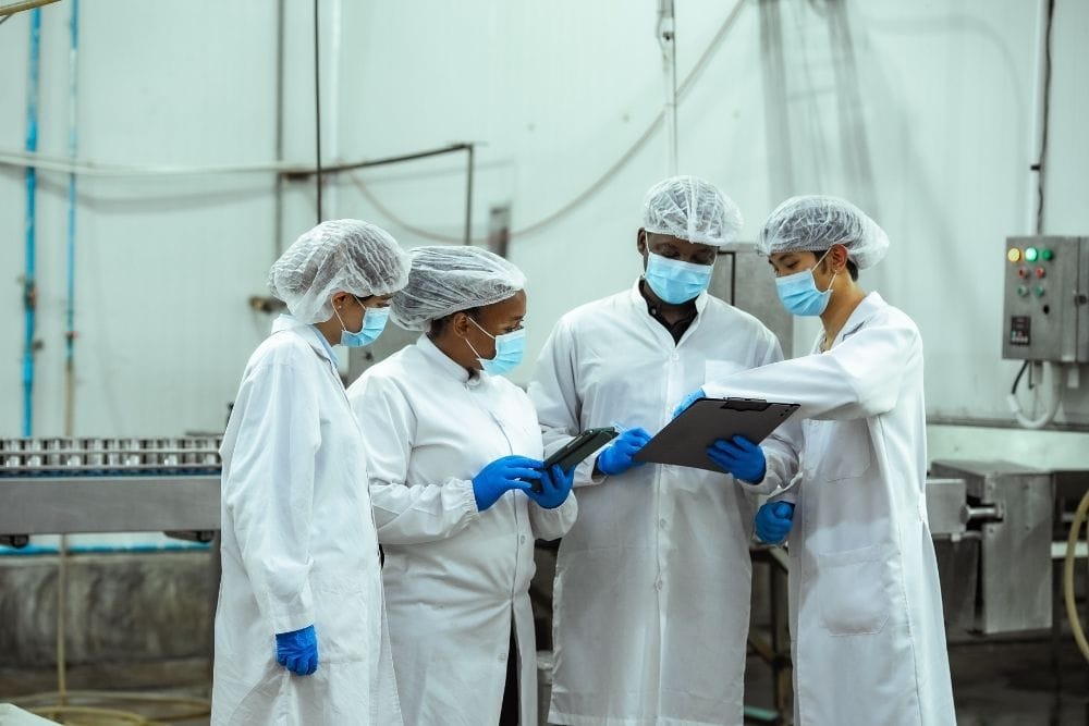 Operator error downtime food production line: Four food production workers in white coats, hairnets, face masks, and blue gloves gathered around a clipboard on the factory floor.