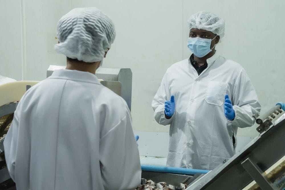 operator knowledge retention food production: A male supervisor in a hairnet, face mask and blue gloves speaking and gesturing to a colleague with their back to the camera on a food production line, with metal machinery in the foreground.